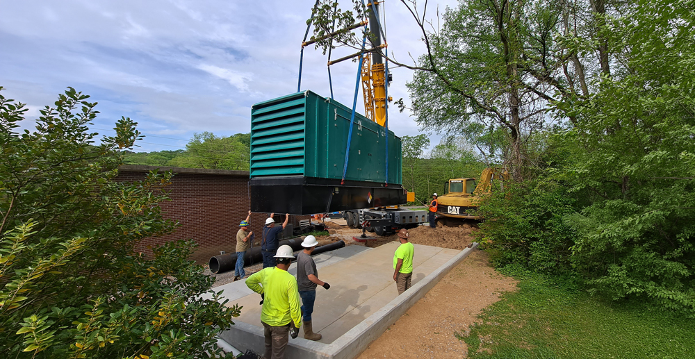 A crane lifting a large diesel generator enclosure onto a concrete pad at a Milcrofton Utility District facility, with construction crew members guiding the installation.